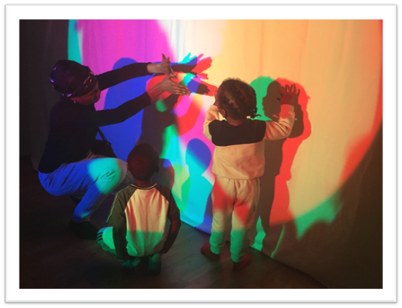 An adult and two children use their hands and bodies to play with coloured lights to cast different coloured shadows and new shapes. No faces visible.