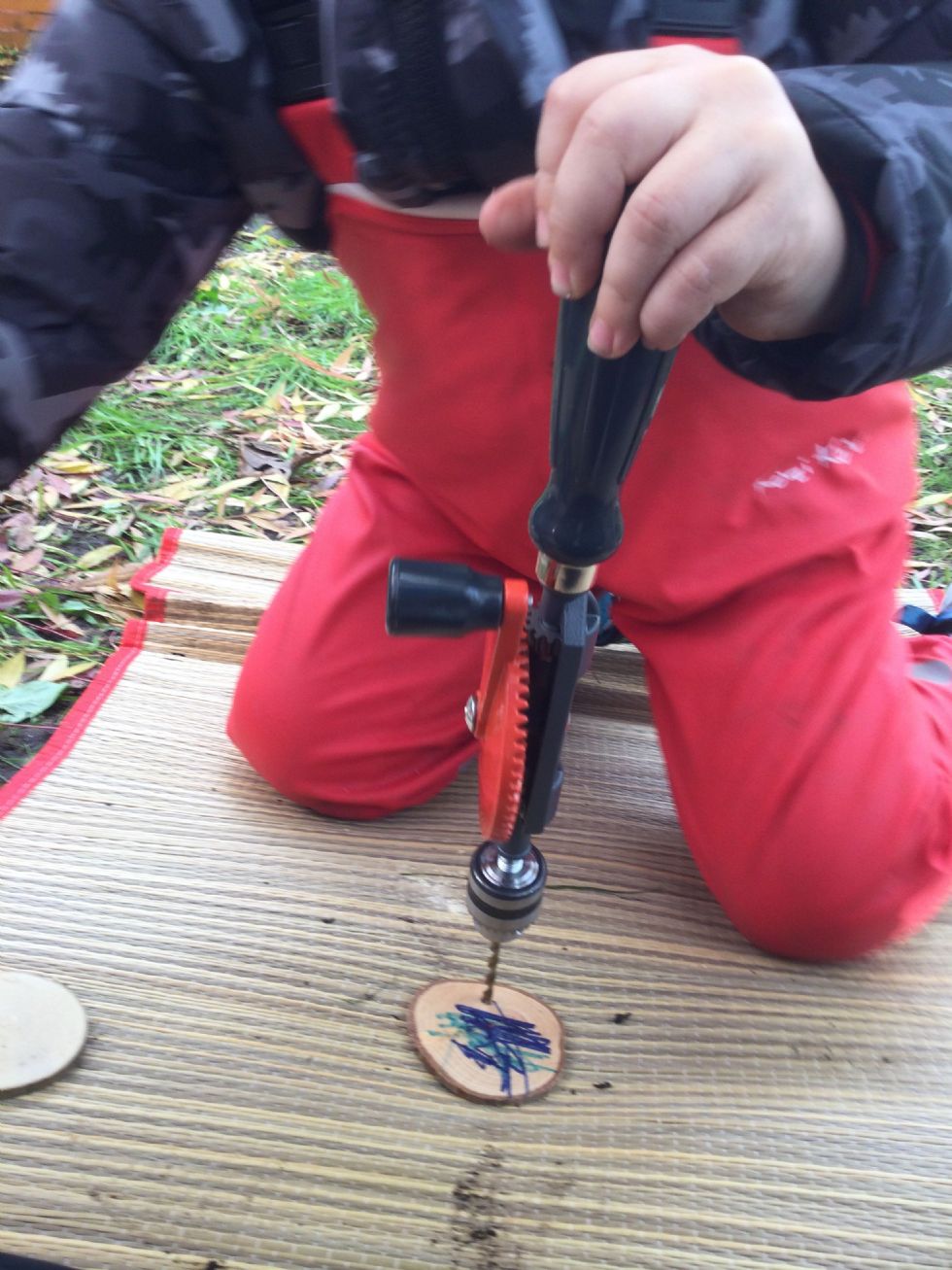 a child wearing waterproof dungarees uses a hand-held drill to make a hole in a piece of wood