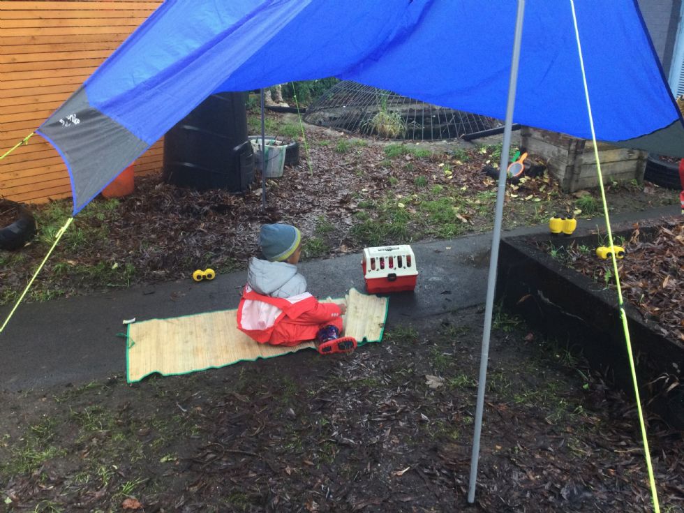 A child wearing a beanie, matching red raincoat and dungarees, and wellies does their work sitting on a rattan mat under a tent sail in the drizzle.