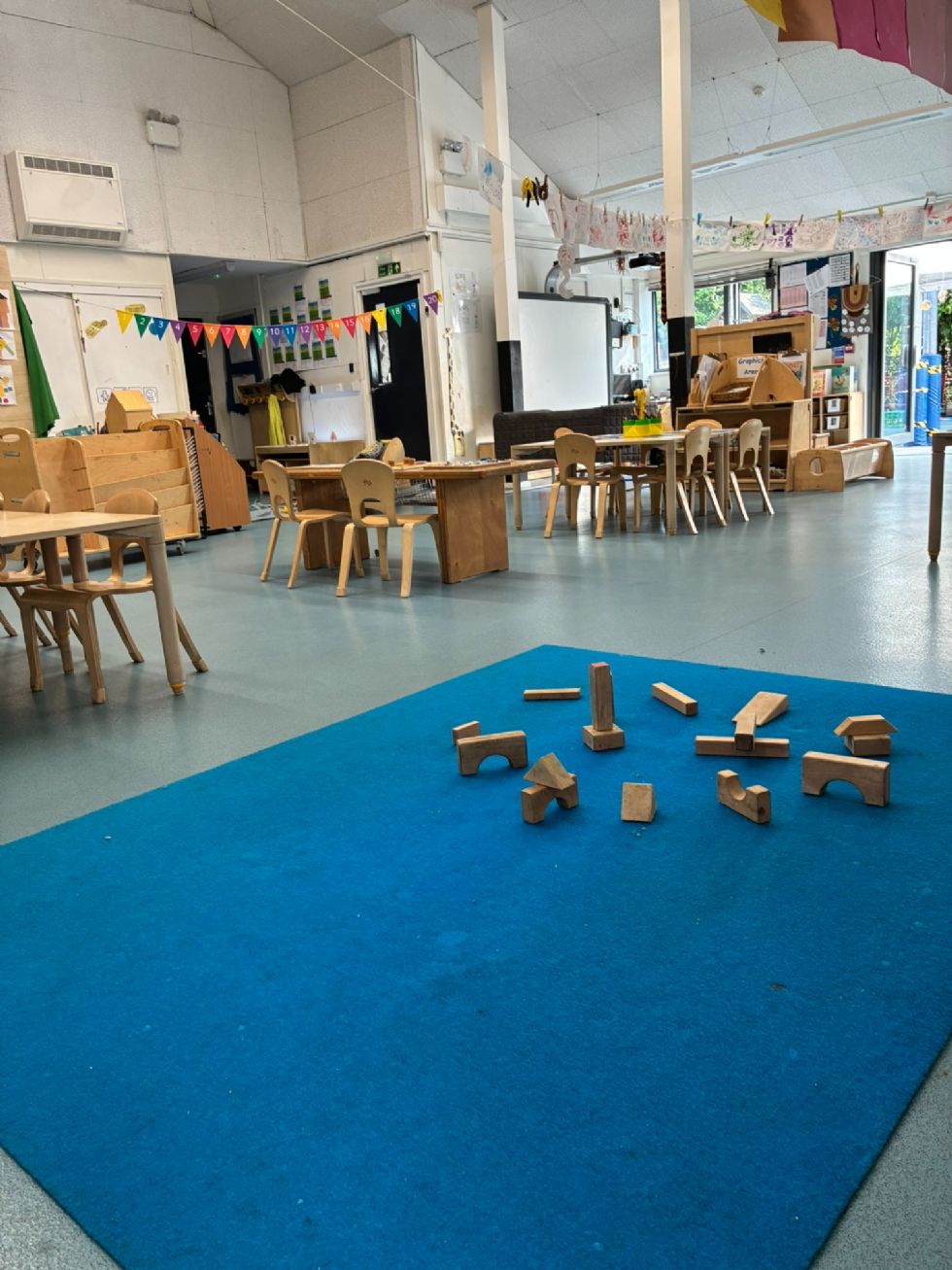 Looking out at the big nursery room with very high ceiling, nearby are blocks, waiting to play. Further off are tables with activities set out. Flags and drawings are strung across the room. In the distance are book shelves and an interactive whiteboard. Right at the back is the open door to the garden.