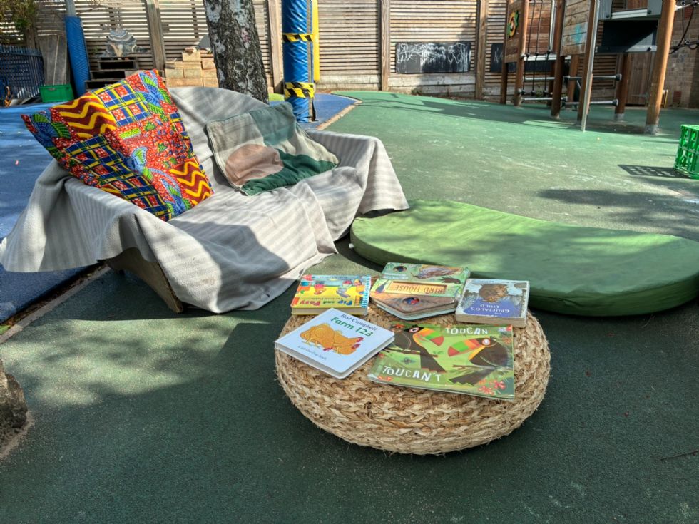The garden has a reading area nearby, with a sofa, cushions, mats, and books. Just behind the sofa is the trunk of one of the silver birch trees which cast shadows on the sunlit scene. In the distance on the right is the bottom of the climbing frame. On the left is a pile of building blocks. 