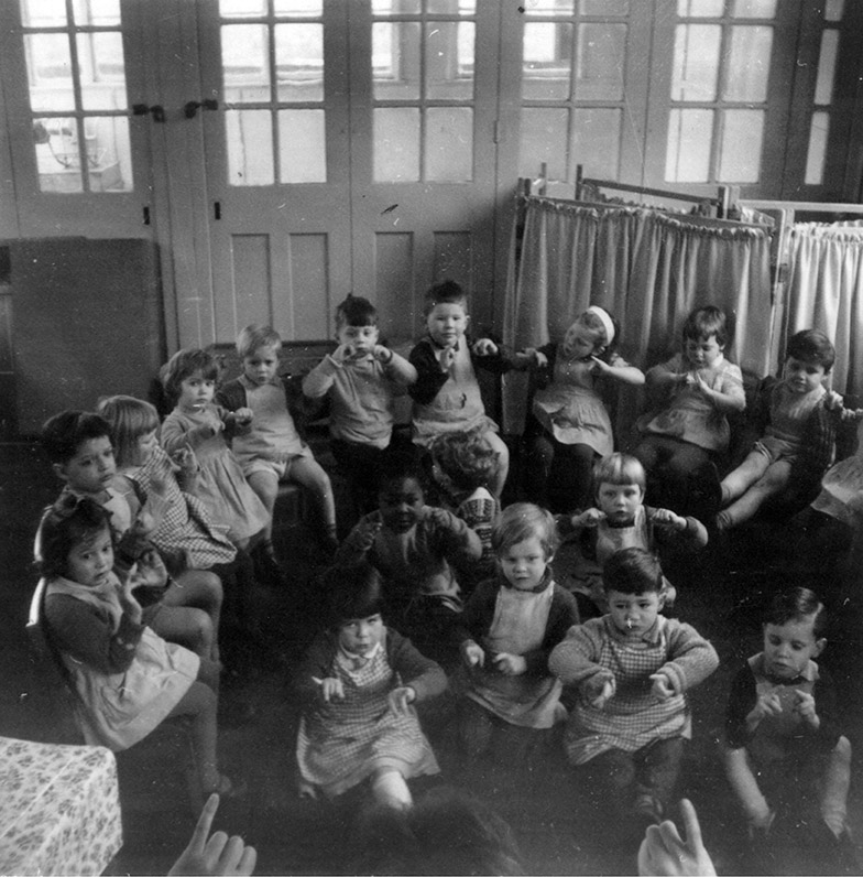 Low Hall Nursery School, c.1963, courtesy Lesley Anne Finlayson black and white photo of 3 to 4 year-old children at Low Hall in c1963, sitting on chairs and on the floor making hand gestures as if miming a song